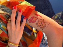 Tatouage temporaire avec un soleil souriant, une flamme et un piment sur le bras d'une personne aux ongles rouges.