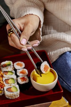 Sushi à emporter servi dans un plateau rouge avec sauce jaune, accompagné de makis au saumon et avocat, main tenant baguettes