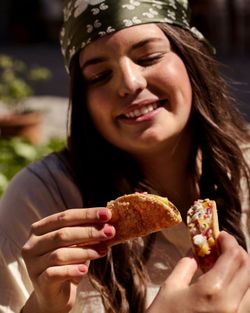 Jeune femme souriante dégustant des tacos, lumière naturelle et décor extérieur flou en arrière-plan.
