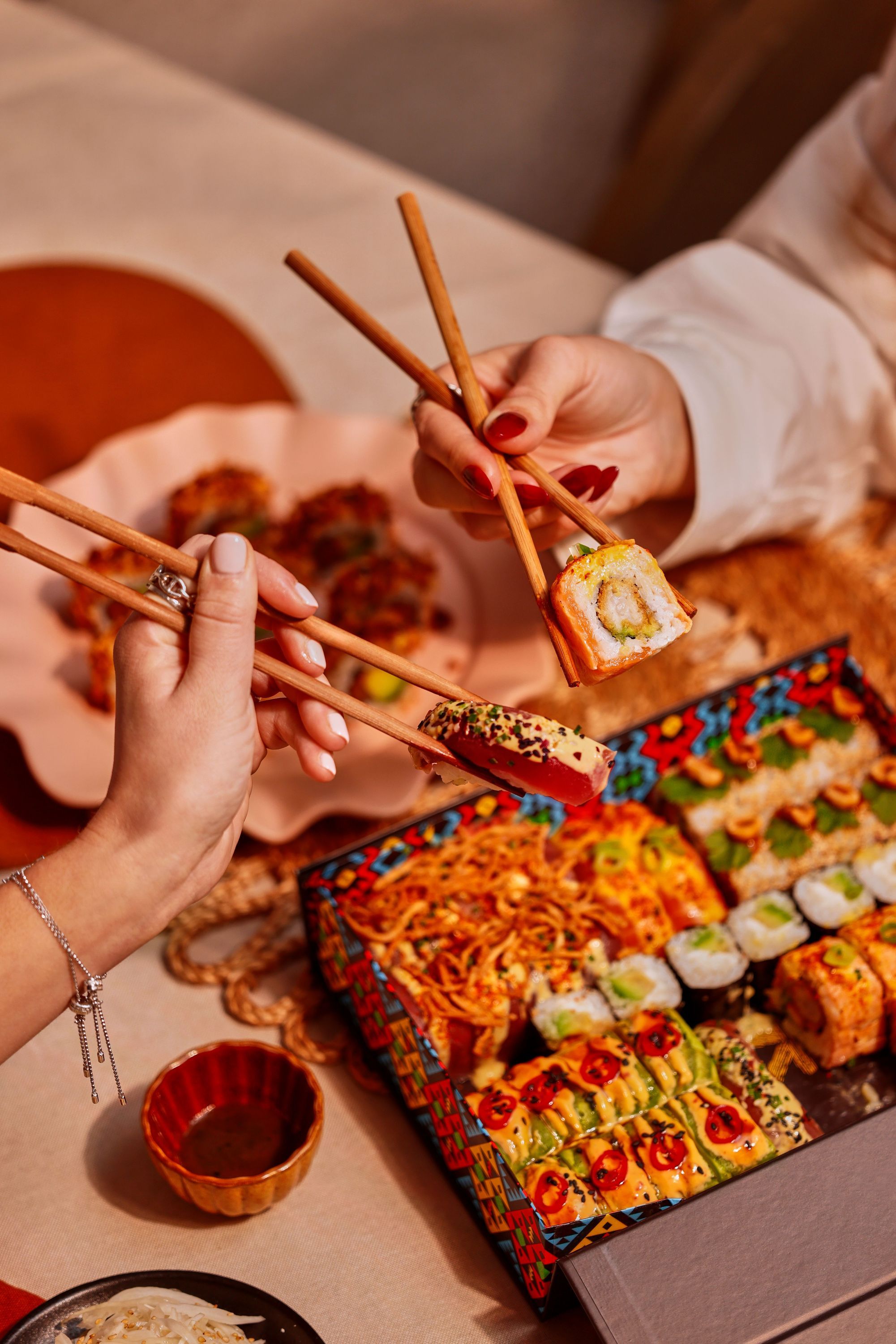 Deux personnes dégustent des sushis avec des baguettes, plateau coloré varié de sushi à emporter visible sur la table.