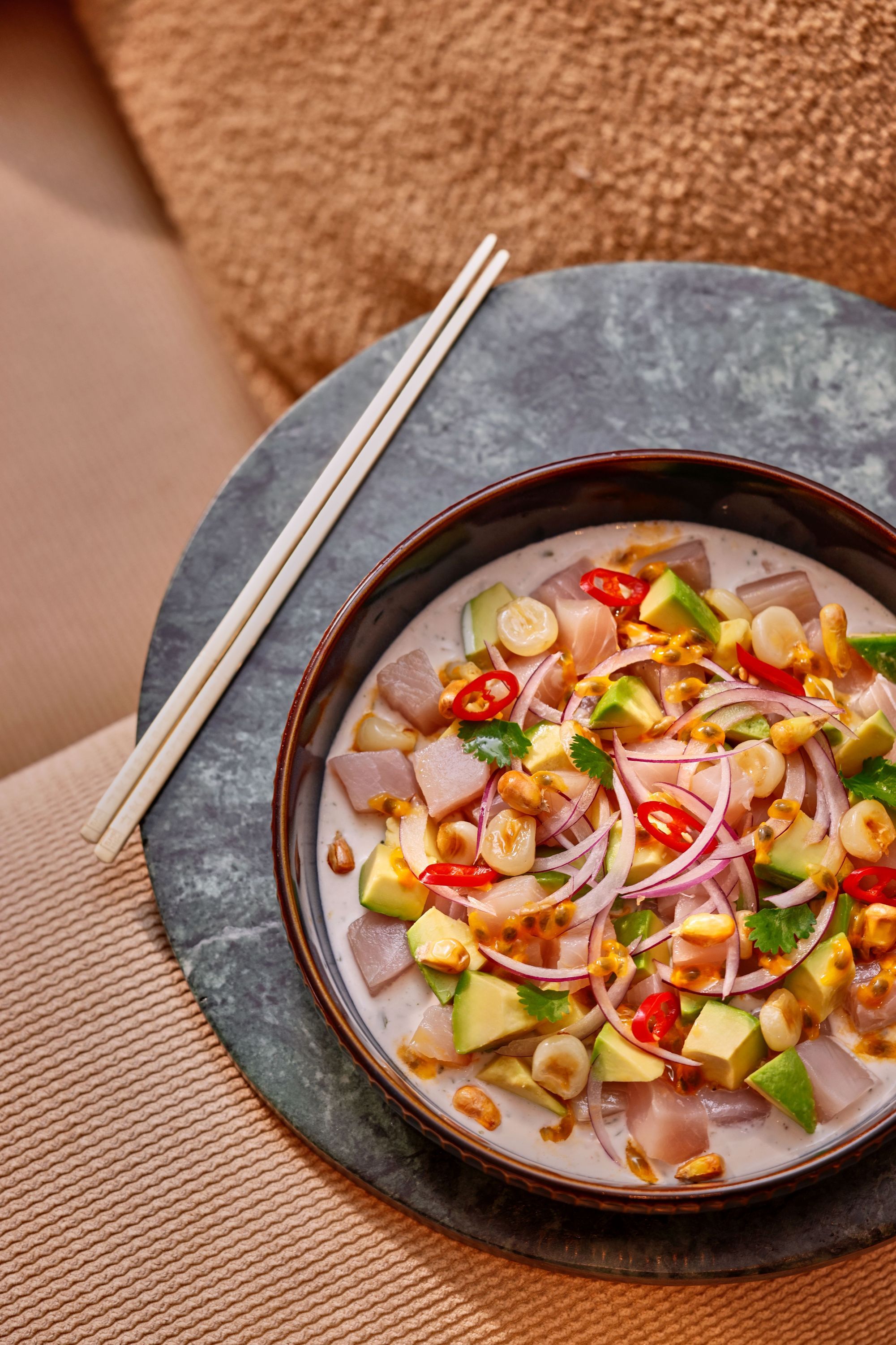 Bol de ceviche avec avocat, oignons, piments et maïs toasté, accompagné de baguettes sur une table ronde en pierre.