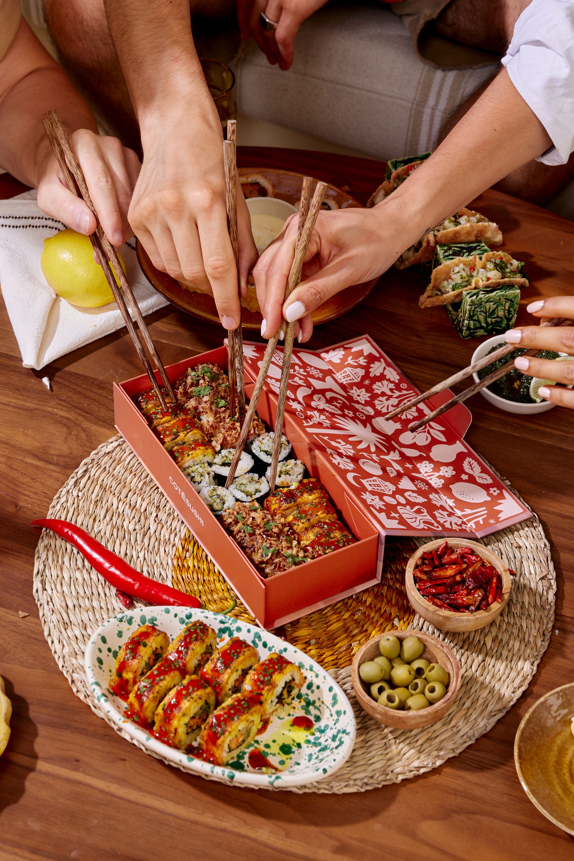 Plusieurs mains utilisent des baguettes pour prendre des sushis dans une boîte rouge sur une table avec des plats variés