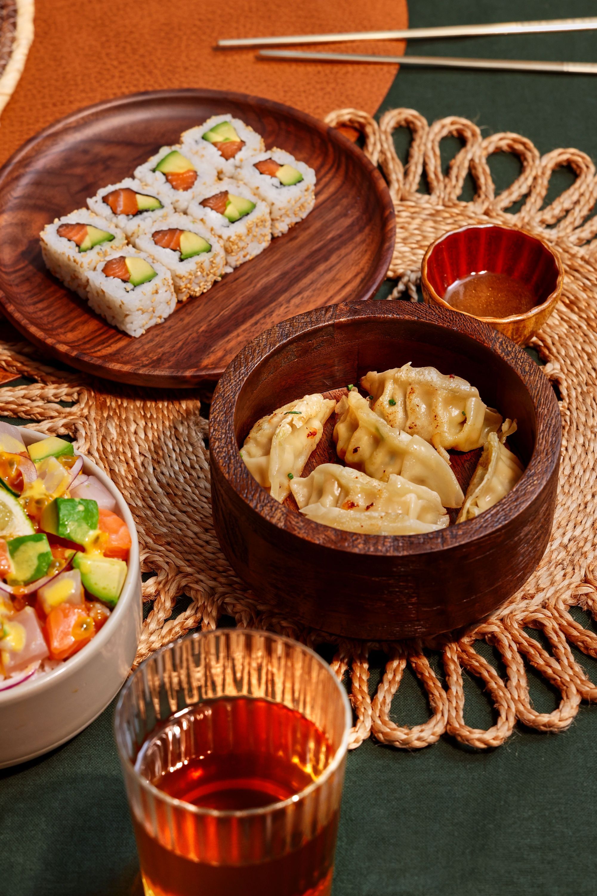 Assortiment de sushis et raviolis japonais dans des bols en bois sur une table avec boisson et sauces.