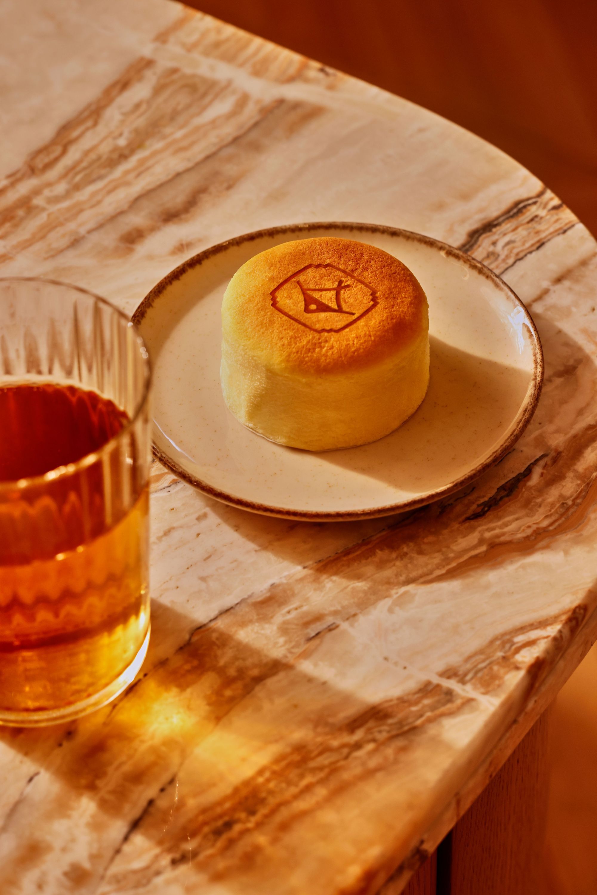Petit gâteau japonais avec logo sur assiette blanche, posé sur une table en marbre clair, à côté d'un verre de boisson.